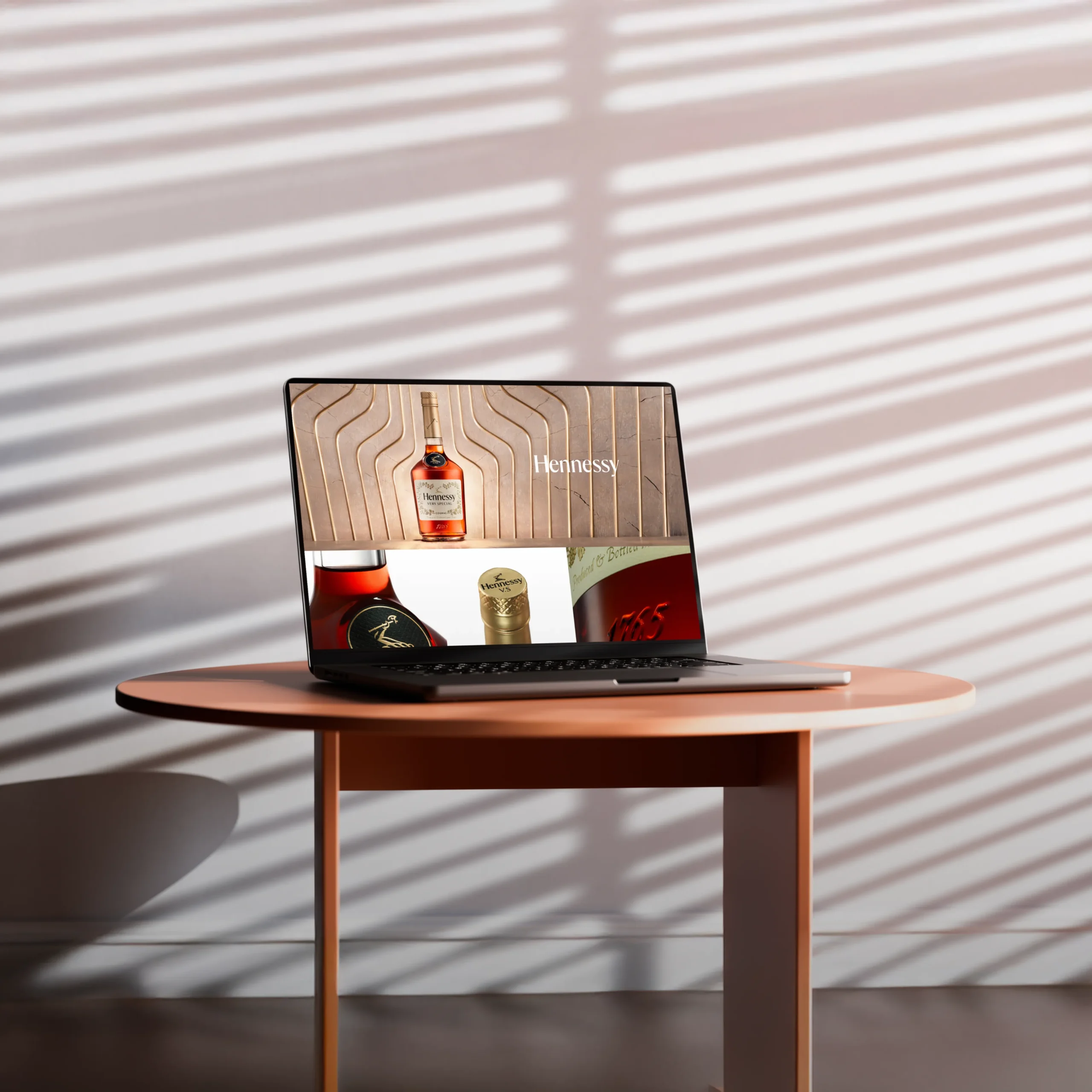 Superchouette- Un ordinateur portable posé sur une table ronde rose affiche une publicité pour le cognac Hennessy. La lumière du soleil filtre à travers les stores, projetant des ombres rayées sur le mur et la table.