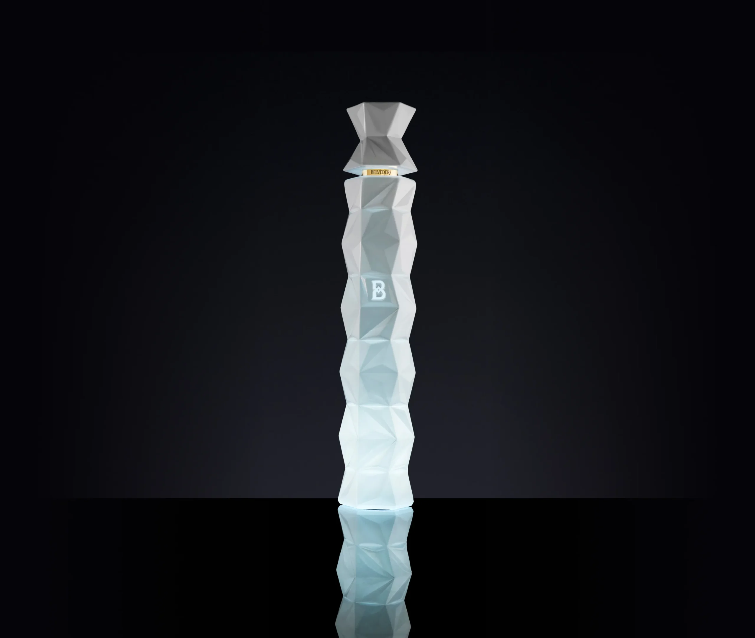 Superchouette- Une grande bouteille géométrique en verre dépoli à facettes avec un B sur sa surface, debout sur une surface noire réfléchissante sur un fond sombre.
