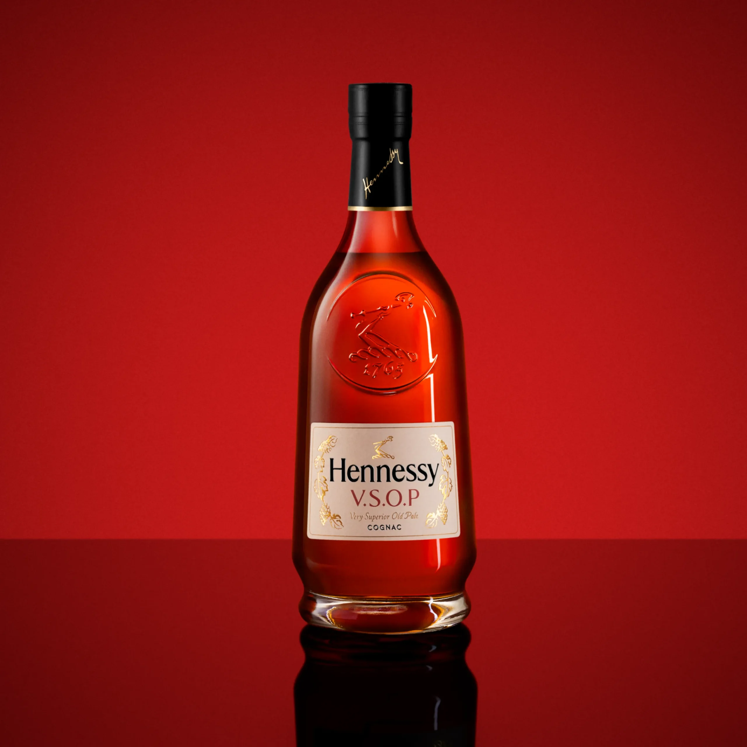 Superchouette- Une bouteille de cognac Hennessy V.S.O.P se tient debout sur une surface réfléchissante à fond rouge, mettant en valeur son étiquette or et blanche et son bouchon sombre.