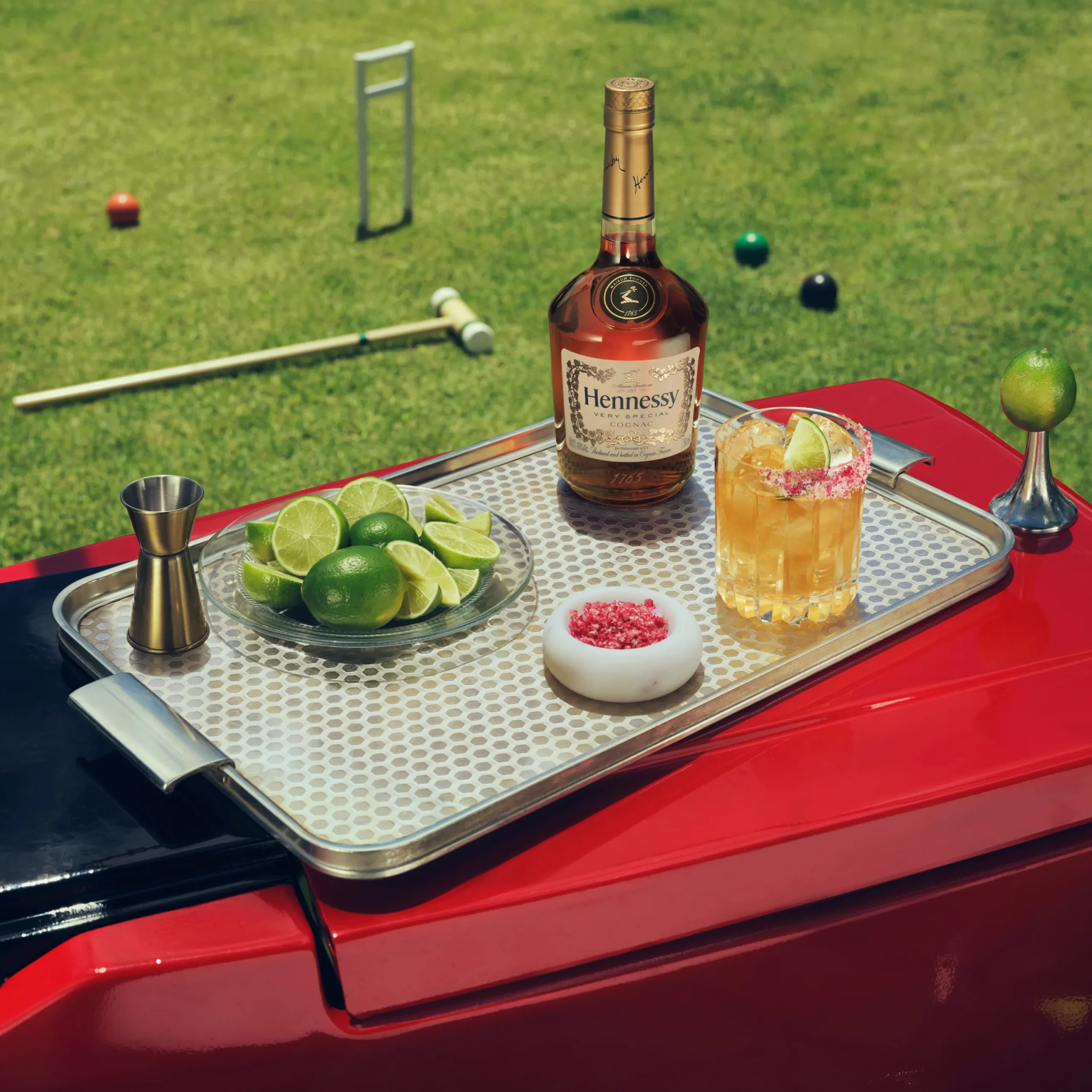 Superchouette- Un plateau métallique avec une bouteille de Hennessy, un cocktail avec des tranches de citron vert, un bol de sel rouge, des quartiers de citron vert, un jigger et des citrons verts sur une surface rouge ; un terrain de croquet herbeux se trouve à l'arrière-plan.