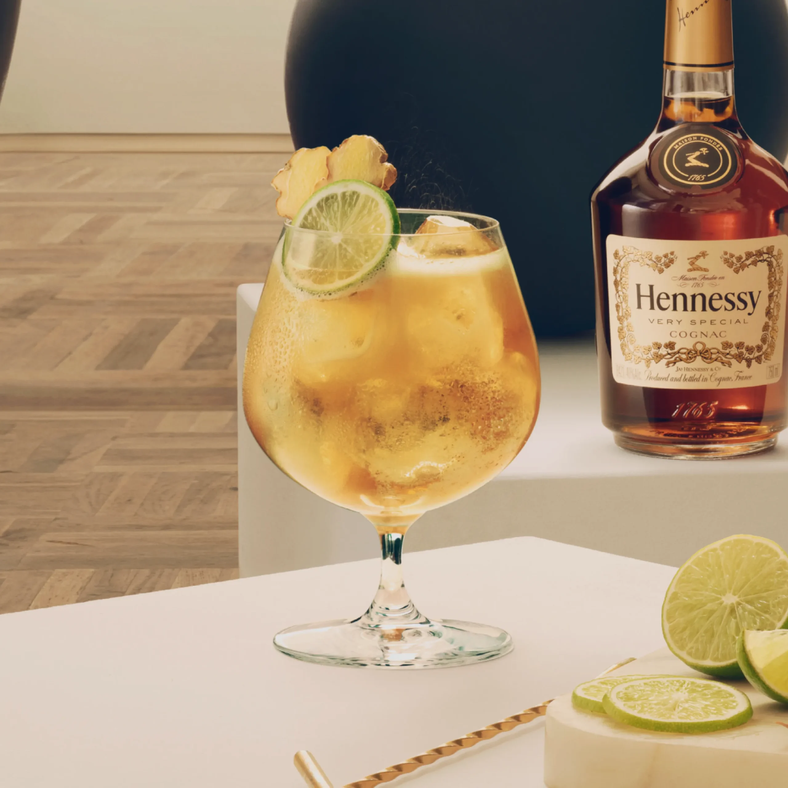 Superchouette- Un verre rempli d'un cocktail glacé garni de tranches de citron vert et de gingembre est posé sur une table à côté d'une bouteille de cognac Hennessy, de tranches de citron vert et d'une paille dorée.