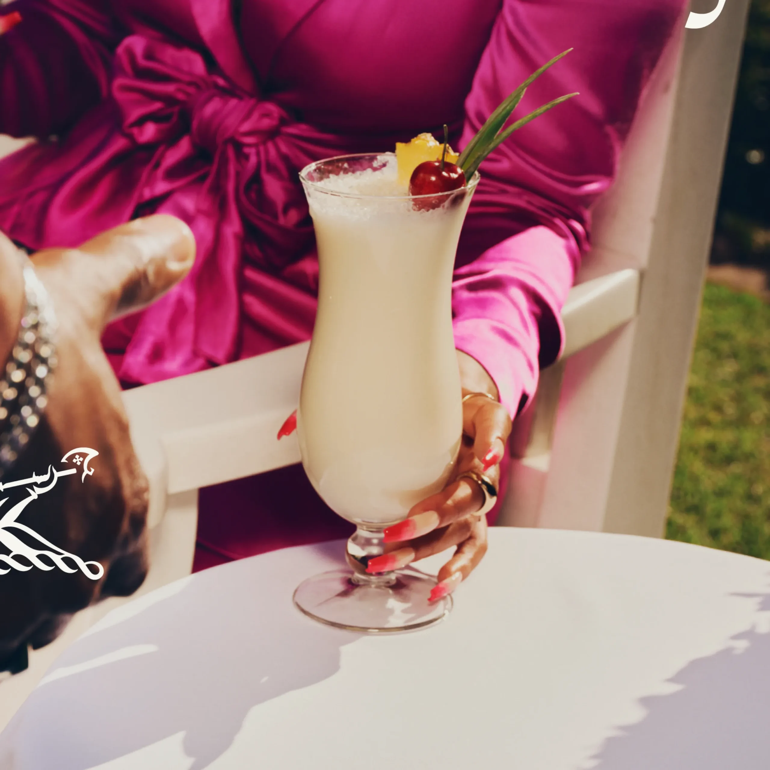 Superchouette- Une personne vêtue d'une robe en satin rose vif tient une pina colada garnie d'ananas, de cerises et d'une feuille, assise à une table blanche en plein air. Une autre main, portant un bracelet, se tend vers la boisson.