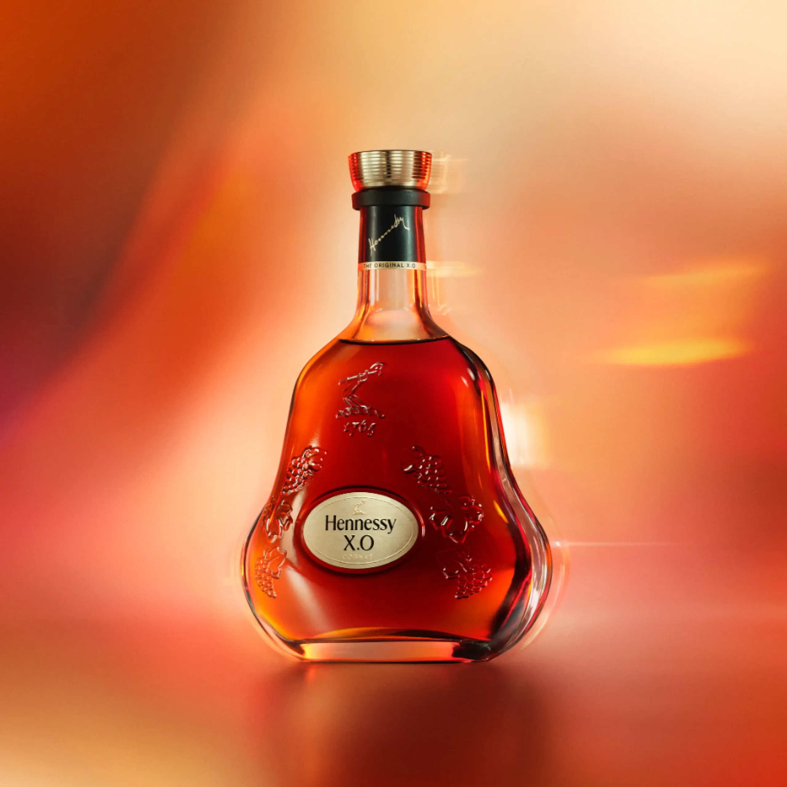 Superchouette- Une bouteille de cognac Hennessy X.O. se tient debout sur un fond orange et or chaud et flou, mettant en valeur les courbes élégantes et les détails en relief de la bouteille.