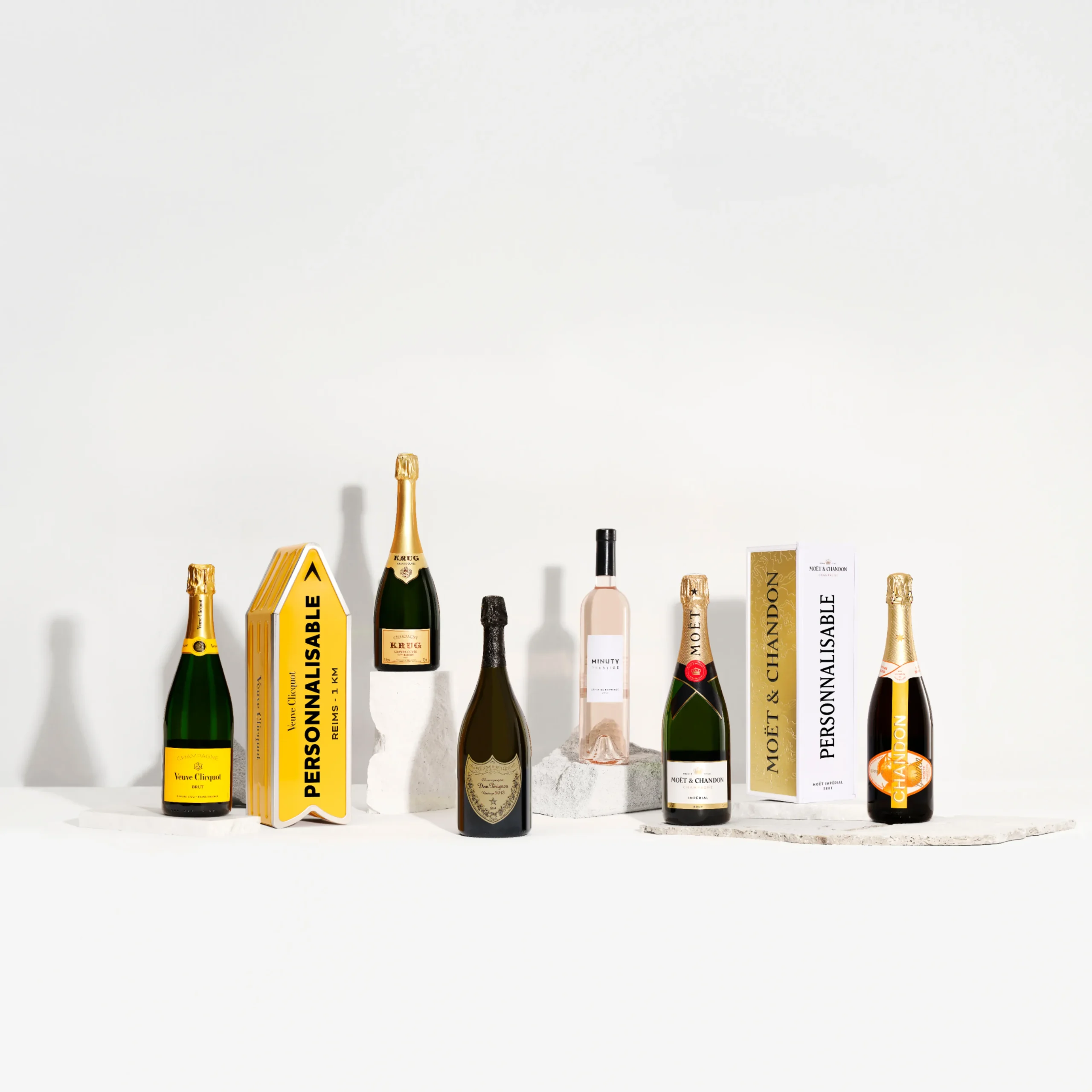 Superchouette- Une sélection de sept bouteilles de champagne et de vin, certaines dans des coffrets cadeaux, disposées sur une surface blanche avec des dalles de pierre pour la présentation sur un fond blanc uni.