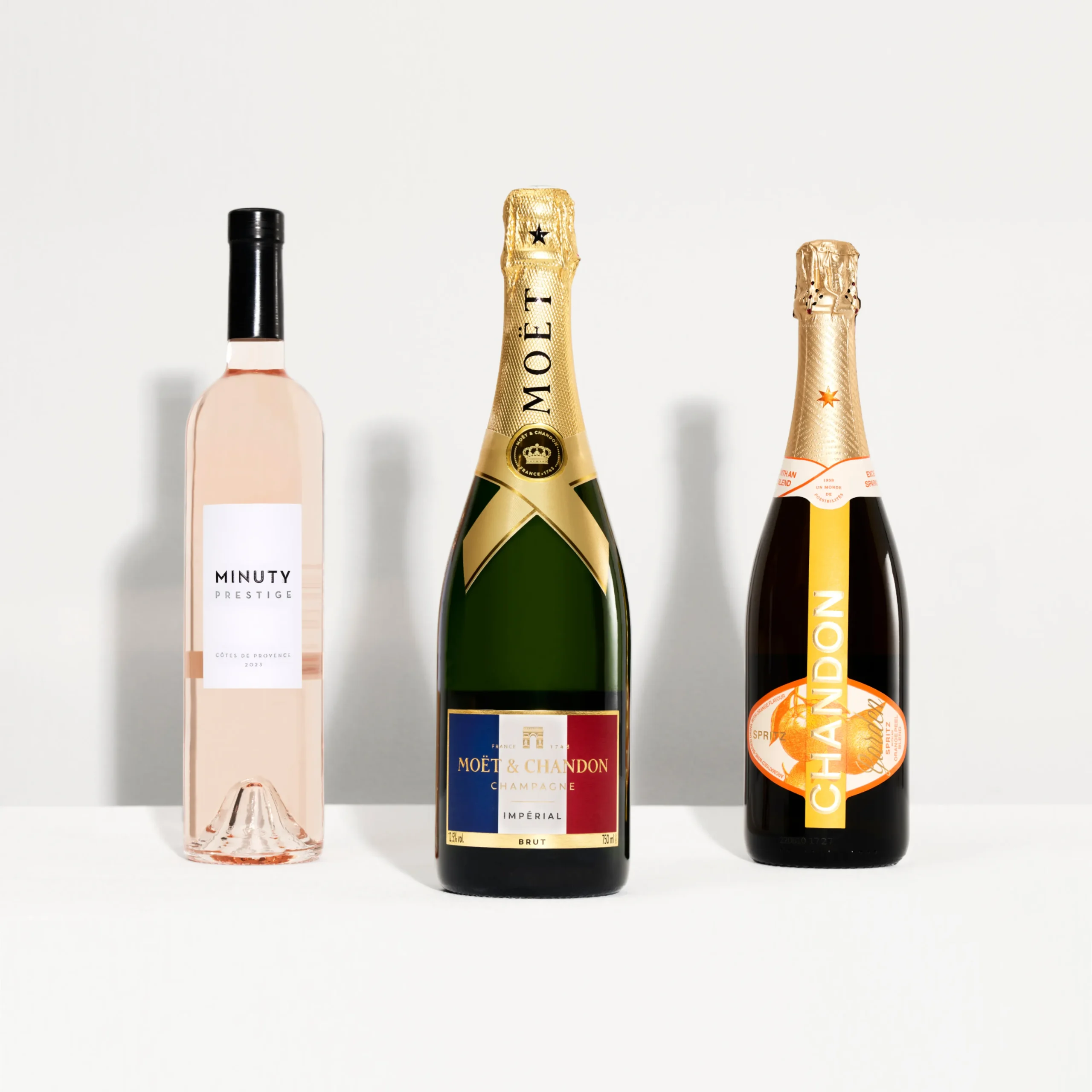 Superchouette- Trois bouteilles de vin mousseux et de rosé sont disposées côte à côte sur une surface blanche : Minuty Prestige rosé (à gauche), champagne Moët & Chandon (au centre) et vin mousseux Chandon (à droite).