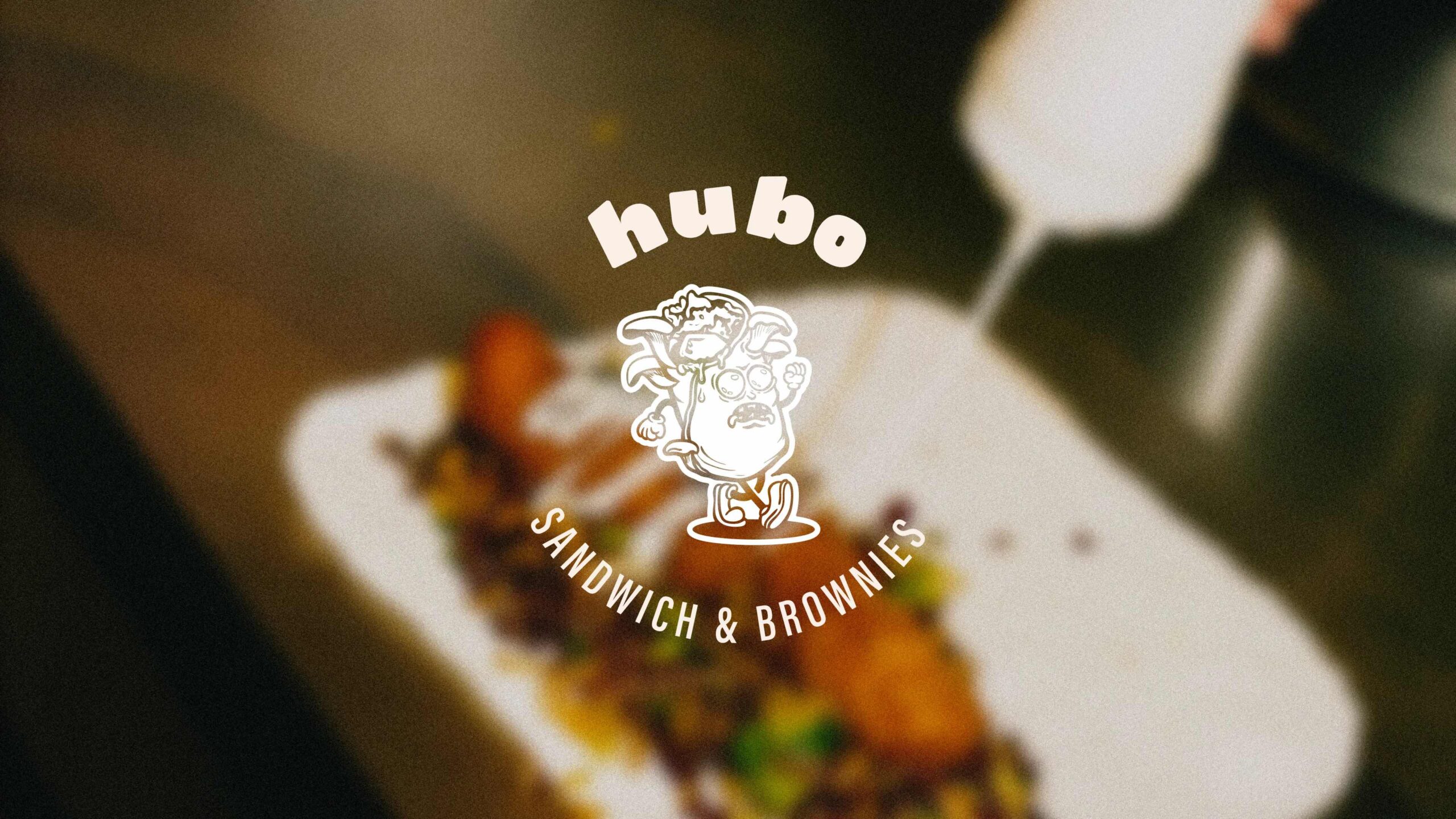 Superchouette- Image floue d'aliments arrosés de sauce, avec un personnage de sandwich en dessin animé et le texte "hubo SANDWICH & BROWNIES" superposé au centre.