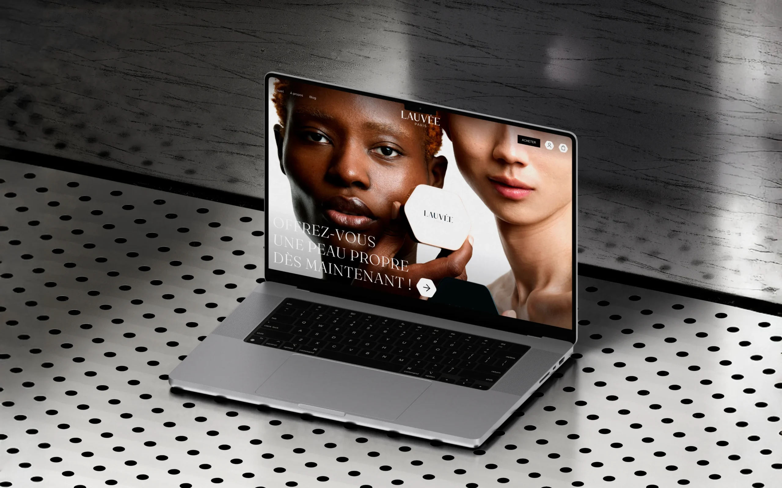 Superchouette- Un ordinateur portable posé sur une surface métallique perforée affiche un site web avec des images en gros plan de deux personnes tenant un produit hexagonal étiqueté Lauvée et un texte en français vantant les mérites d'une peau claire.