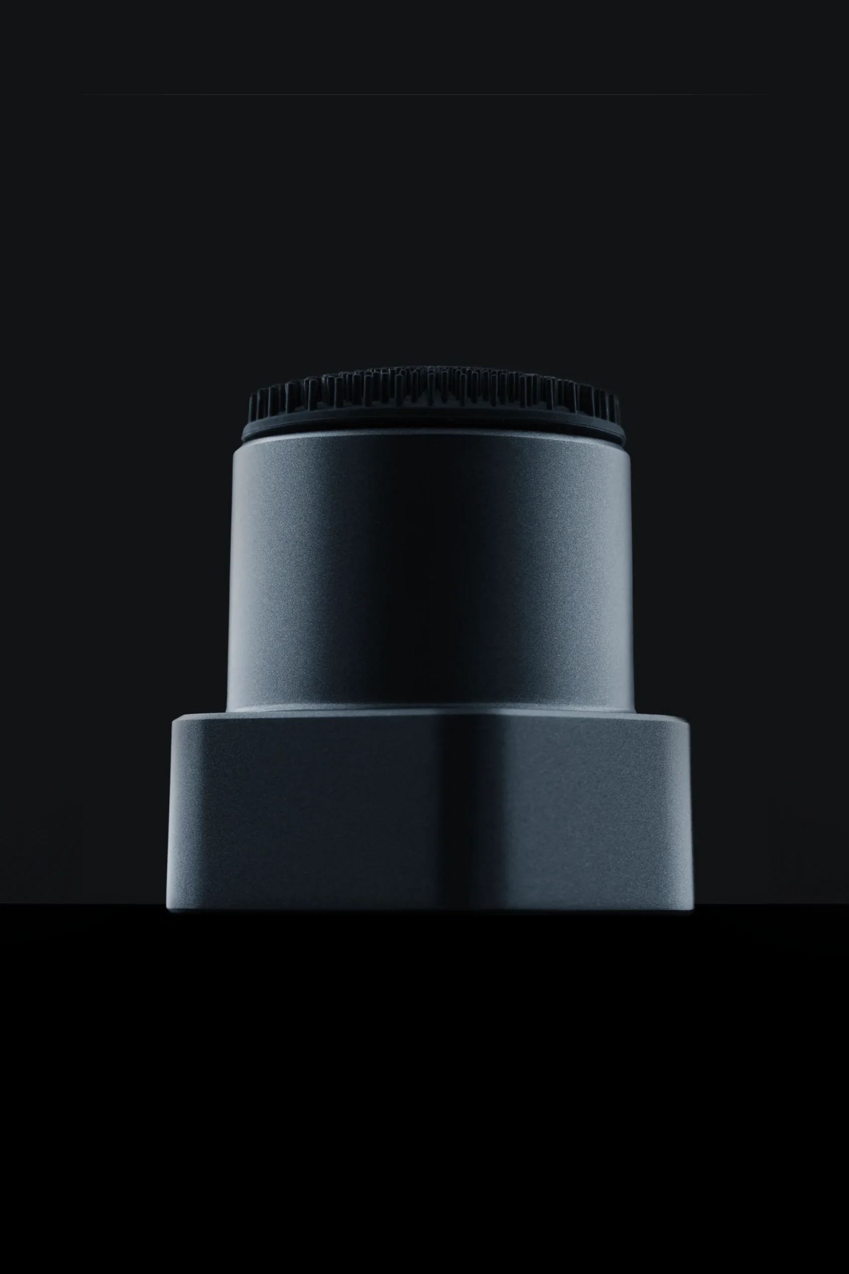 Superchouette- Photo minimaliste aux tons sombres d'un objet métallique cylindrique au sommet texturé, centré sur un fond noir. L'objet semble de conception industrielle ou technologique.