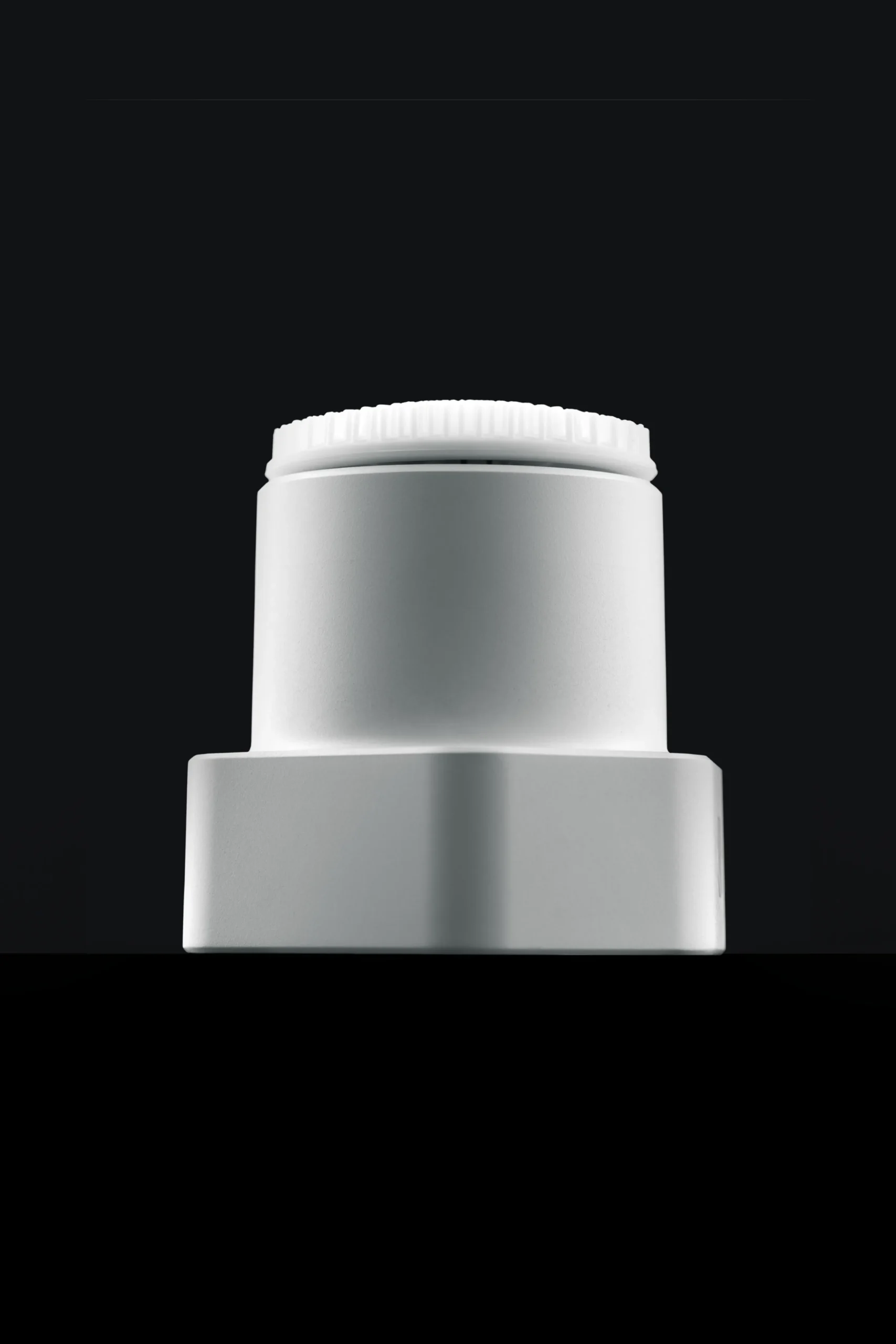 Superchouette- Gros plan sur un bouchon de bouteille en plastique blanc aux bords striés, sur un fond noir uni. Le bouchon est centré et éclairé, ce qui met en évidence sa forme lisse et cylindrique.