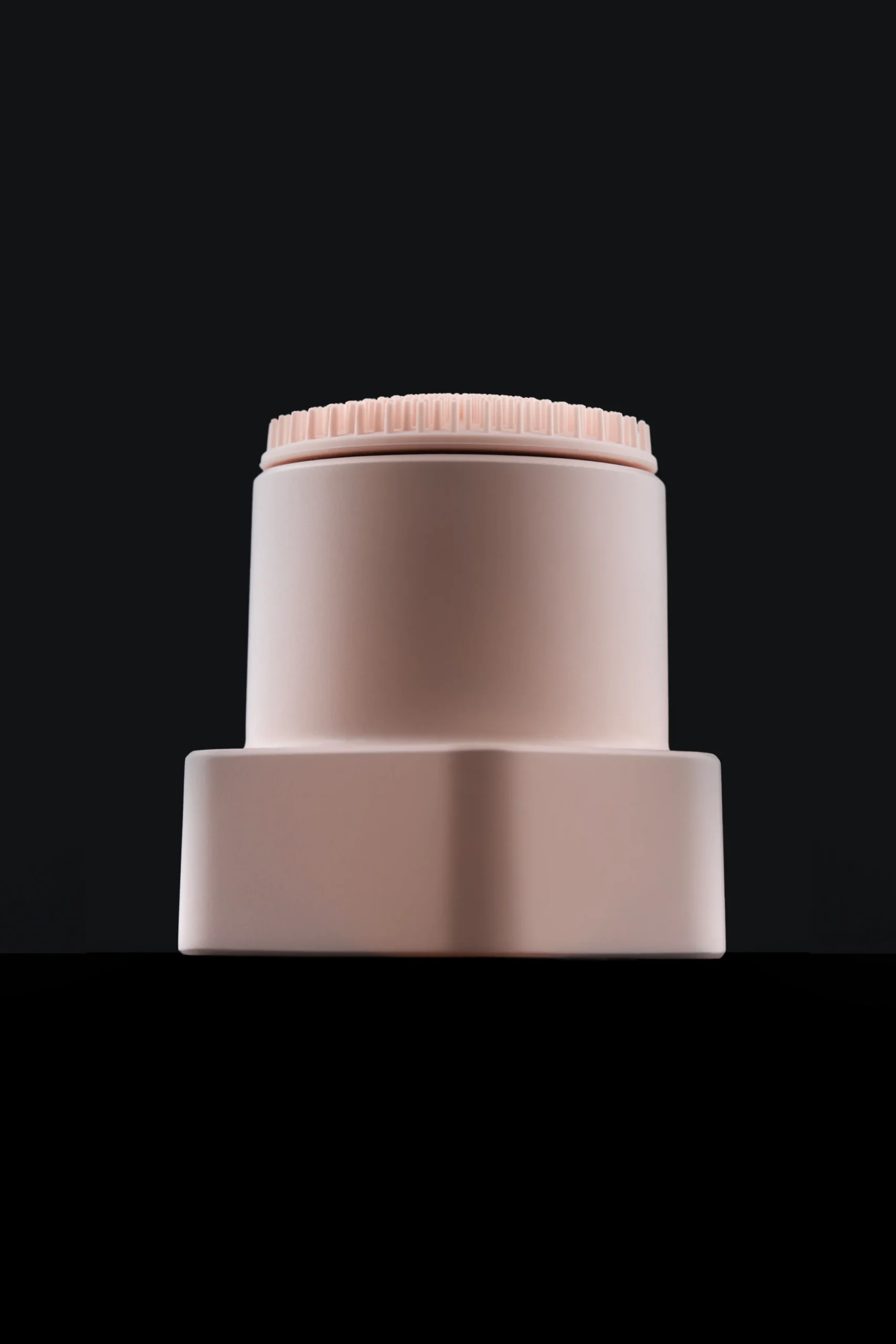 Superchouette- Photo minimaliste d'un récipient cylindrique beige avec un bouchon rose pâle texturé, sur un fond noir uni.