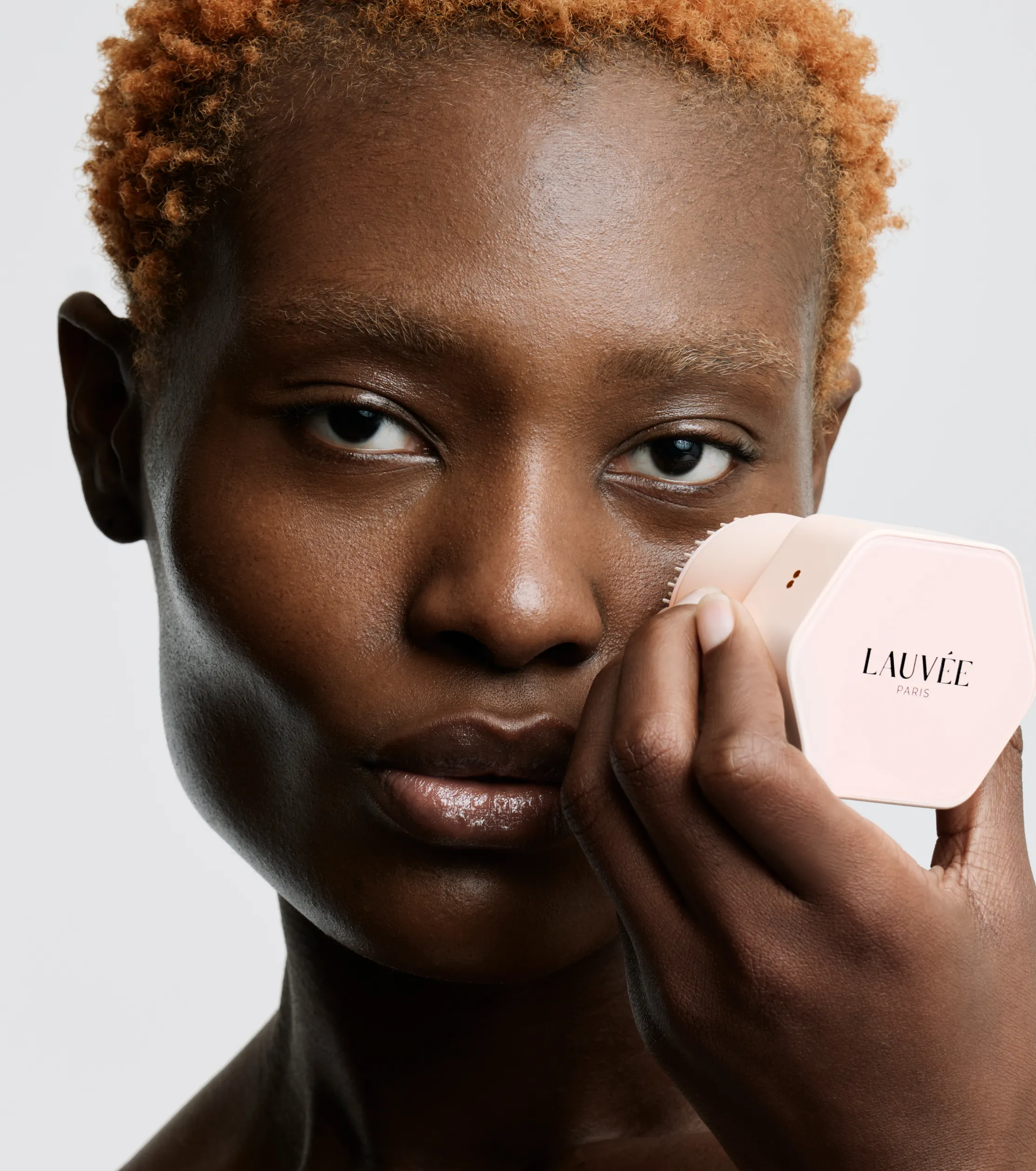 Superchouette- Une personne aux cheveux courts, bouclés et châtains clairs tient un récipient hexagonal étiqueté LAUVÉE PARIS près de son visage, regardant directement la caméra sur un fond gris clair.