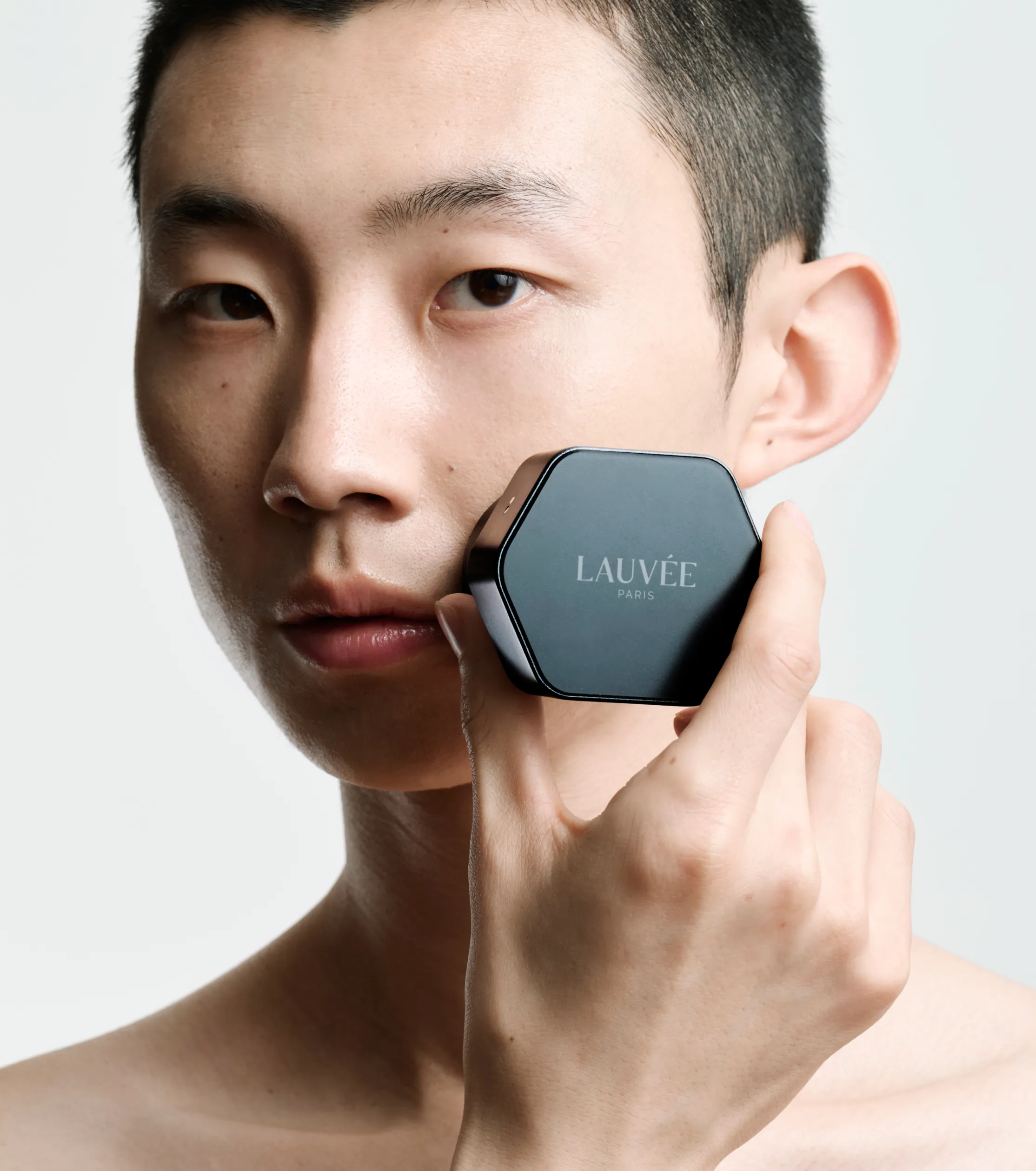 Superchouette- Une personne aux cheveux courts tient sur sa joue un poudrier hexagonal noir étiqueté LAUVÉE PARIS, posant sur un fond clair uni.