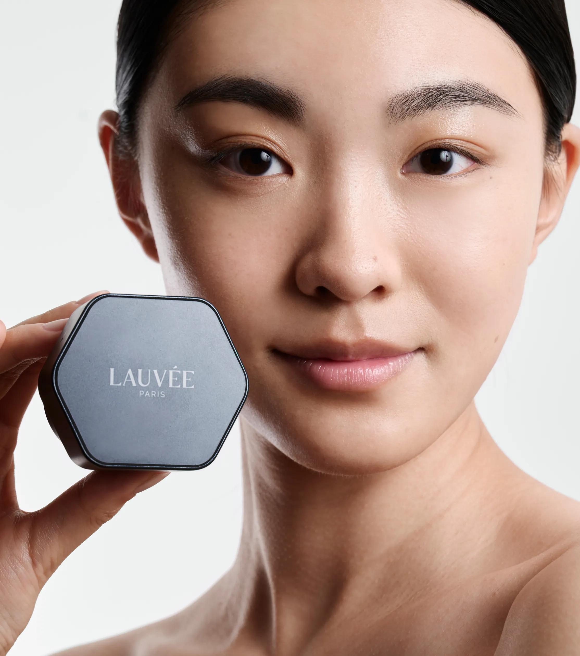 Superchouette- Une femme à la peau lisse tient près de son visage un récipient hexagonal étiqueté LAUVÉE PARIS et sourit doucement sur un fond clair et uni.
