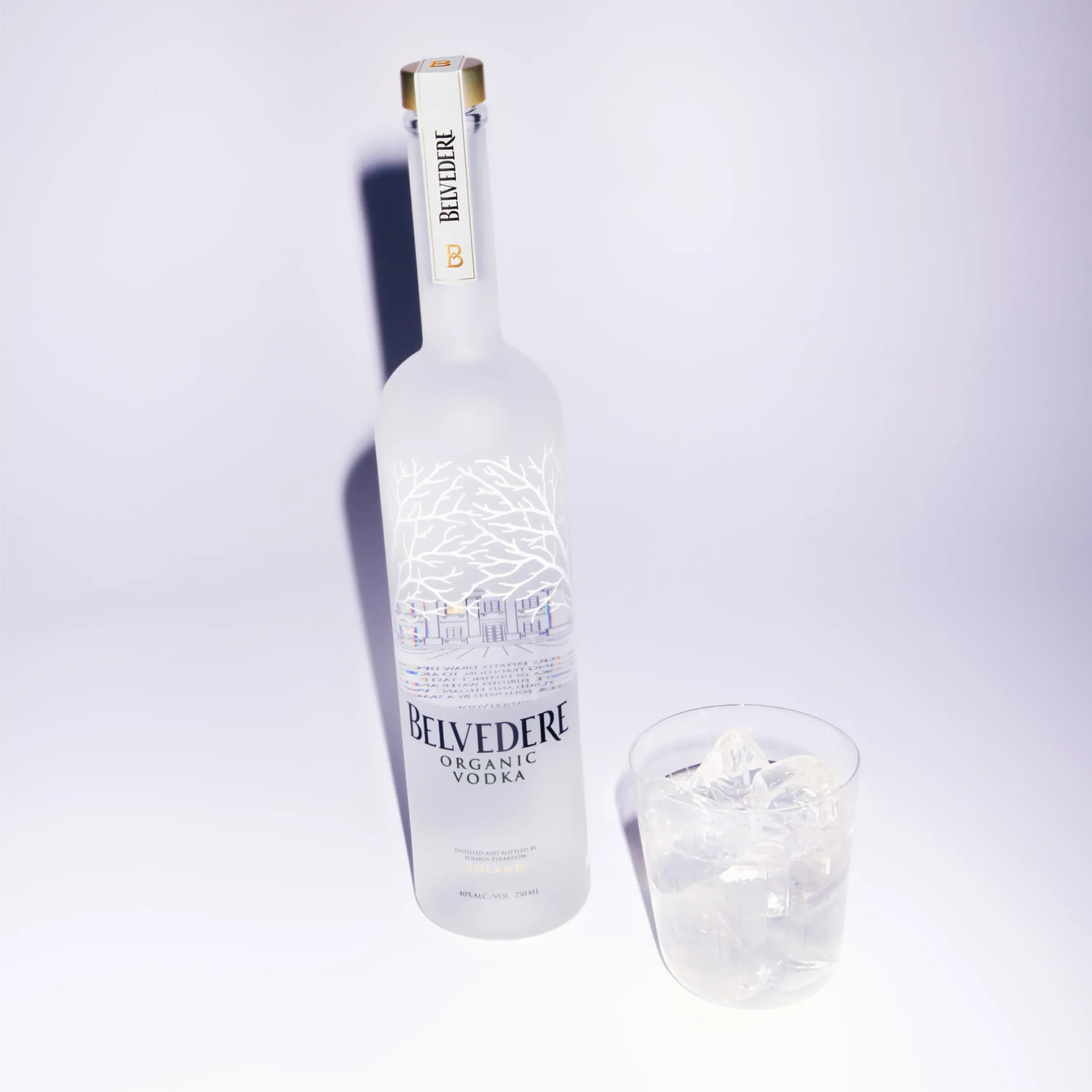 Superchouette- Une bouteille de vodka biologique Belvedere en verre givré se trouve à côté d'un verre rempli de glaçons transparents, le tout sur un fond blanc uni.