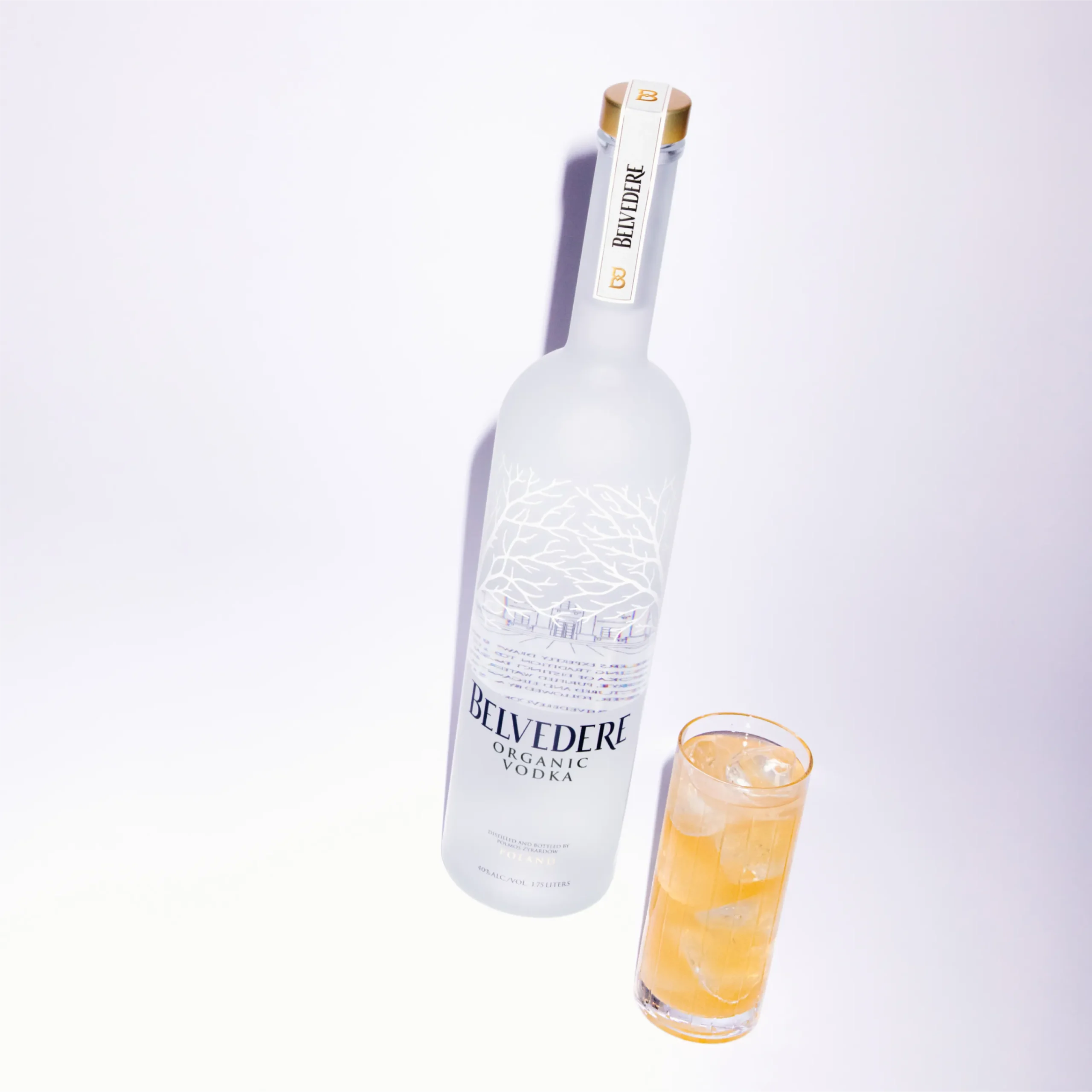 Superchouette- Une bouteille givrée de vodka biologique Belvedere se trouve à côté d'un grand verre rempli d'une boisson légèrement ambrée et de glace, sur fond blanc.