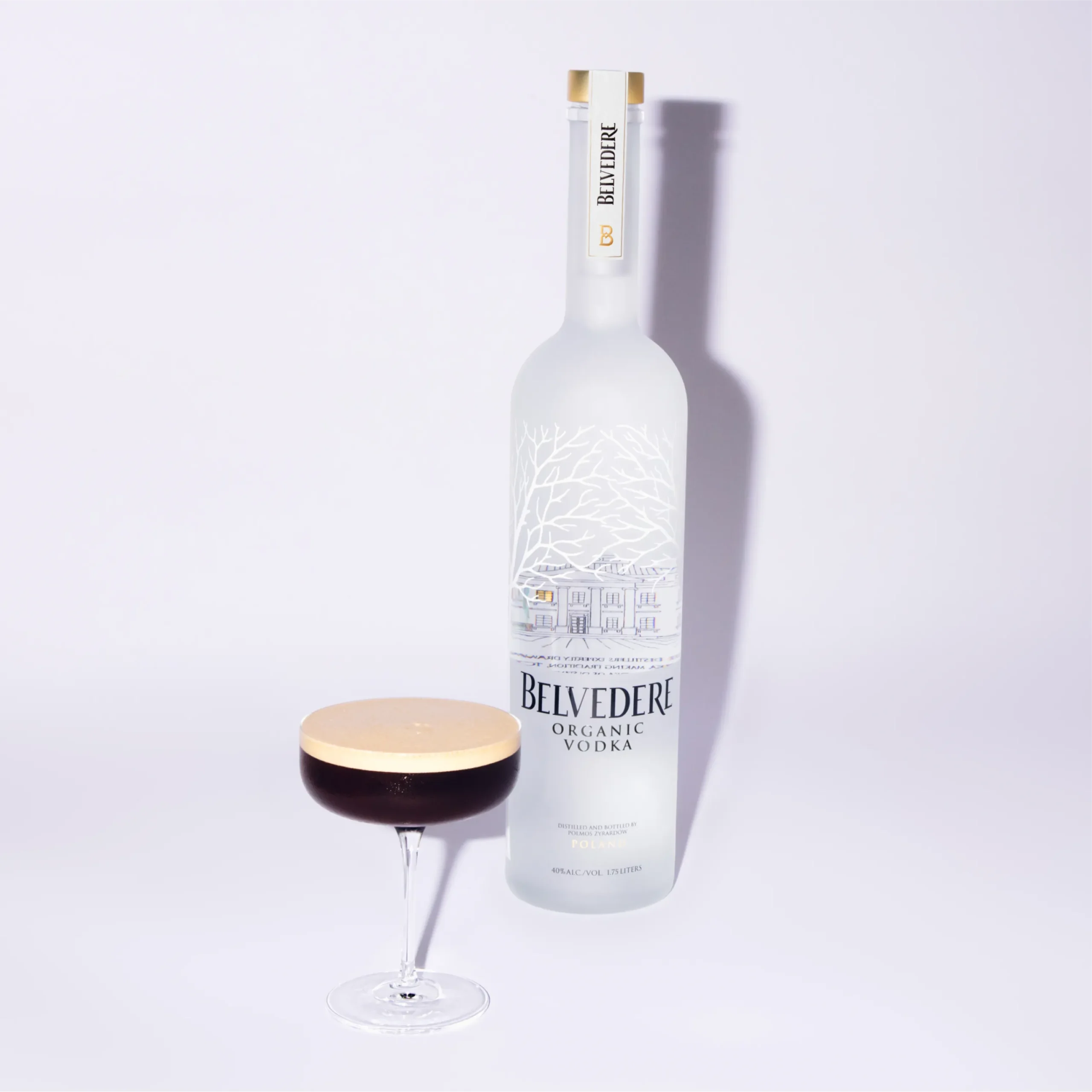 Superchouette- Une grande bouteille de vodka biologique Belvedere se trouve à côté d'un verre à cocktail rempli d'une boisson foncée surmontée d'une mousse crémeuse, le tout sur un fond blanc uni.