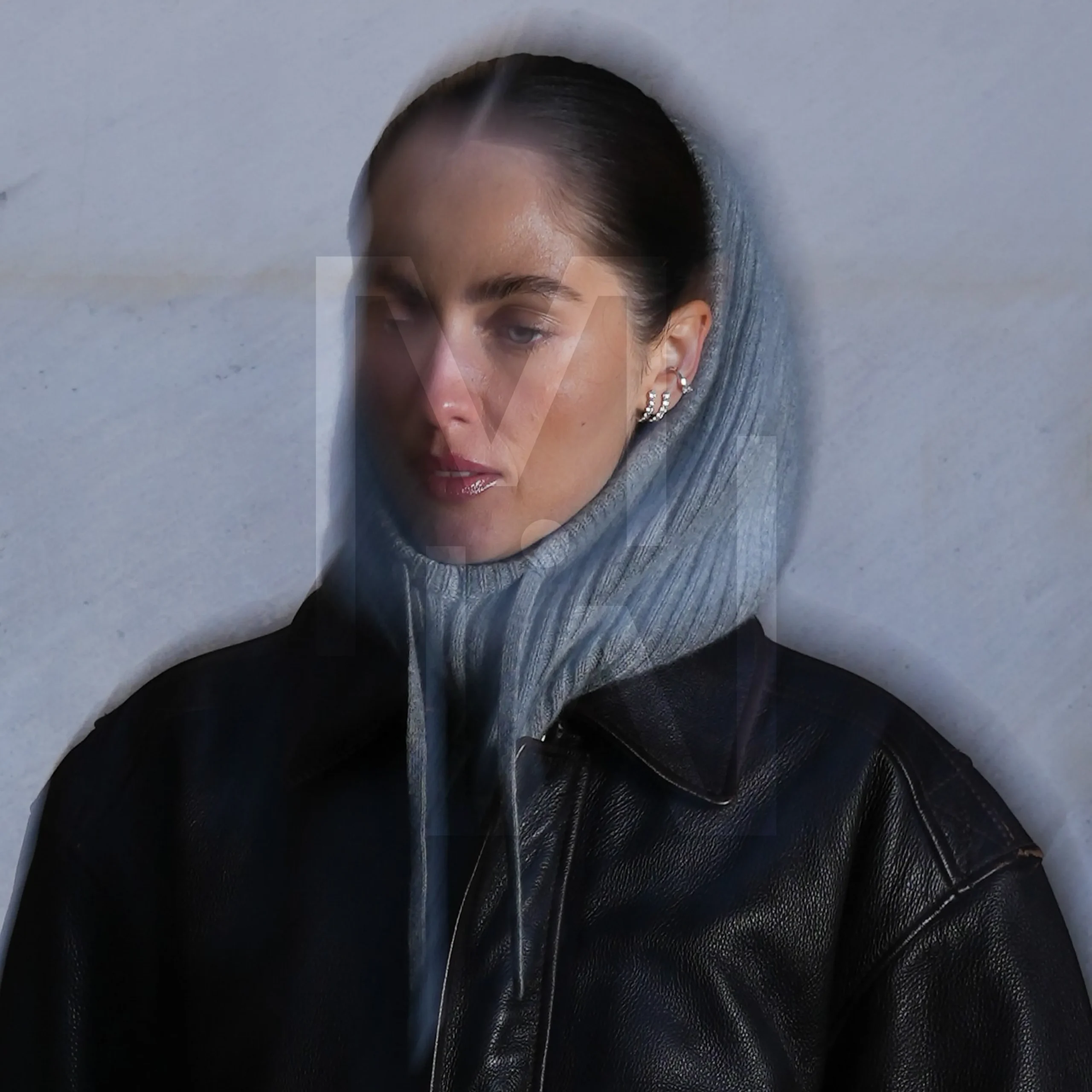 Superchouette- Une femme vêtue d'un sweat à capuche gris et d'une veste en cuir noir se tient contre un mur gris clair, avec un effet de double exposition floue sur son visage et le haut de son corps.