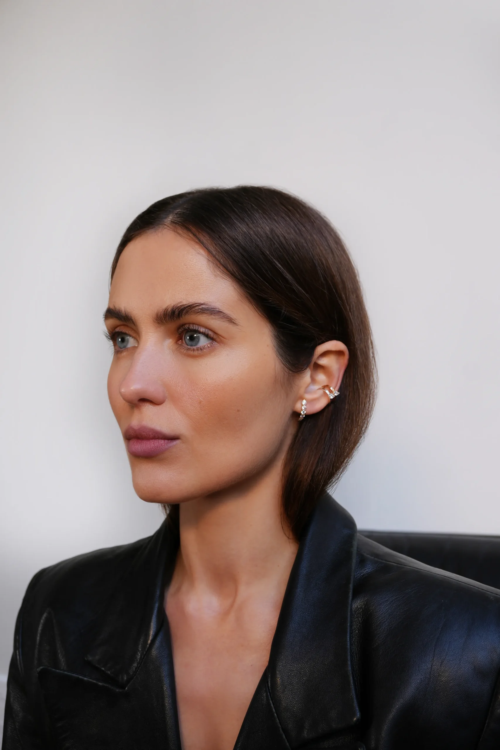 Superchouette- Une femme aux cheveux bruns raides et aux yeux bleus porte une veste en cuir noir et une boucle d'oreille en or. Elle regarde légèrement de côté sur un fond clair et uni.
