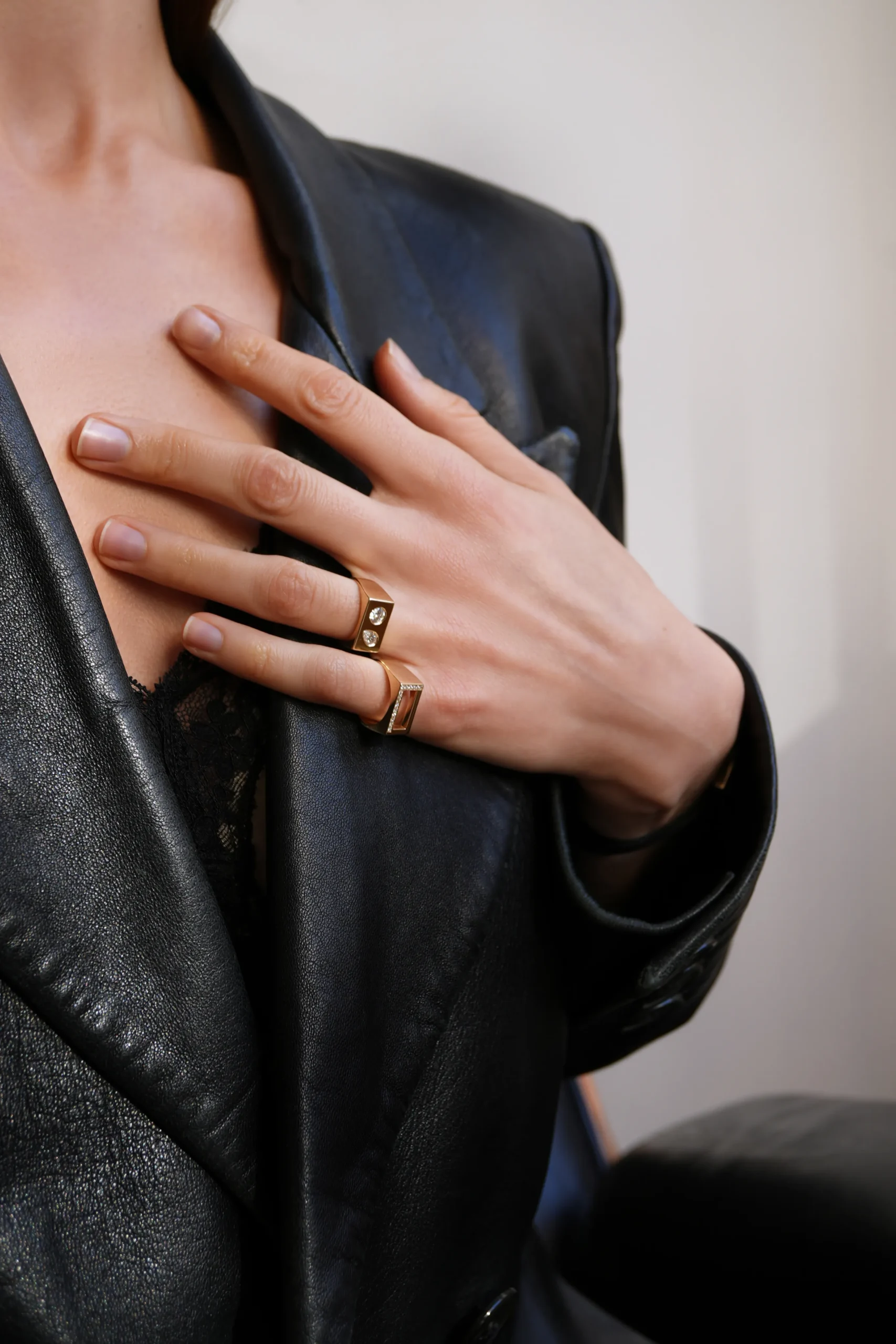 Superchouette- Une personne portant un blazer en cuir noir et un haut en dentelle se touche la poitrine d'une main, montrant deux bagues en or à ses doigts.